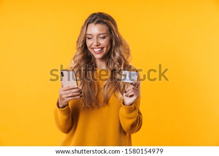 Image of beautiful blonde woman with long hair wearing warm sweater holding credit card and cellphone isolated over yellow background