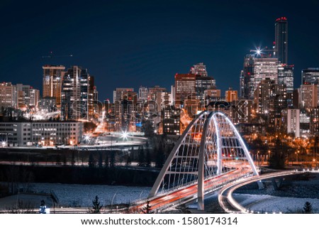 Light trails on the Walterdale Bridge, in Edmonton, Alberta. The lit up city skyline is visible in the background and the sky is a dark blue. Blue and Orange tones. Royalty-Free Stock Photo #1580174314
