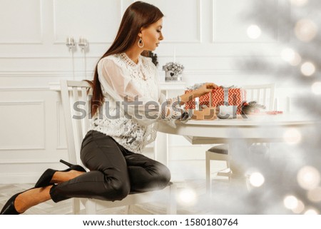 Slim young woman in home interior packing christmas gifts.Free space for your decoration.Xmas tree and white walls. Blurred lights and sun light from window.Copy space. 
