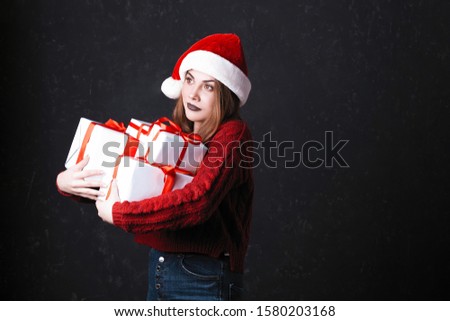 Lovely girl with make-up in a burgundy sweater and santa claus hat. Posing on a dark background holds gifts on a Christmas tree, the concept of Christmas and New Year