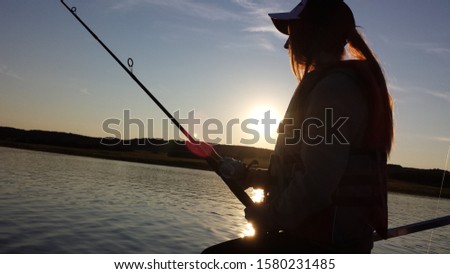 Silhouette of young girl fishing, holding rod on the lake with the sun setting behind her.