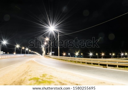 The night highway is illuminated by bright lights and shutter speeds. Factory with tailpipes. The moon and stars, clouds and green grass. The inscription in Russian: "Astrakhan".