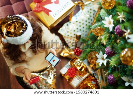 Stylish housewife with long brunette hair in gold sequin skirt and white sweater with credit card making donation via smartphone application under decorated Christmas tree near present boxes.