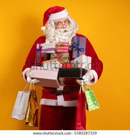 Santa Claus with a long beard and glasses holds a lot of gift boxes and packages in his hands and poses on a yellow background