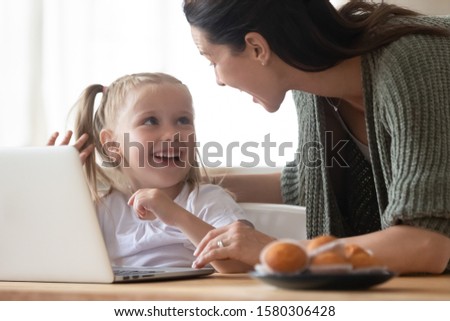Happy mother and little daughter having fun with laptop close up, laughing at funny news, playing video game or watching cartoons, shopping online, smiling mum teaching adorable child to use computer