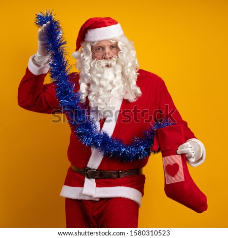 Santa Claus with a long beard and glasses holds in his hands a Christmas sock with gifts and a blue garland of tinsel and poses on a yellow background