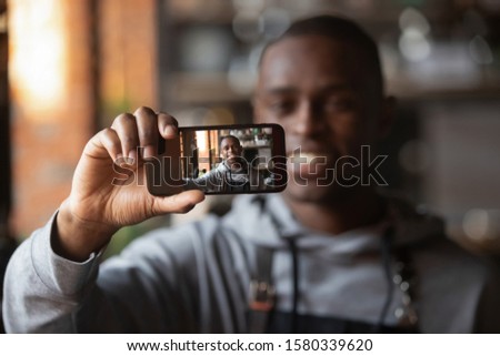 Smiling african American millennial waiter wearing black apron holding smartphone taking selfie in cafe, happy positive black male staff member make self-portrait picture on cellphone in restaurant
