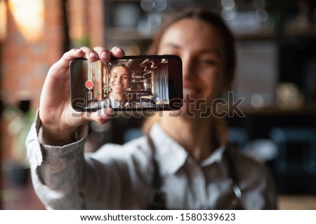 Happy young waitress vlogger holding smartphone recording video blog on mobile display, smiling millennial cafe owner coffeehouse worker blogger girl wear apron shooting vlog looking at phone camera Royalty-Free Stock Photo #1580339623