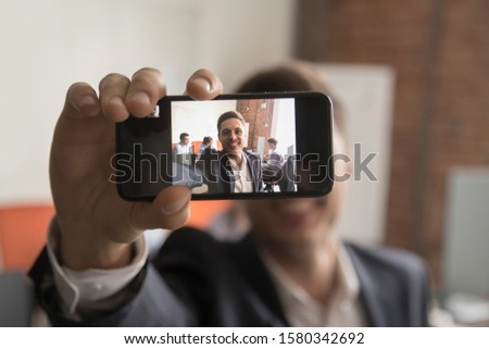 Smiling Caucasian millennial male worker hold smartphone look at camera making self-portrait picture, happy positive businessman extend hand take selfie on cellphone posing in shared office foreground
