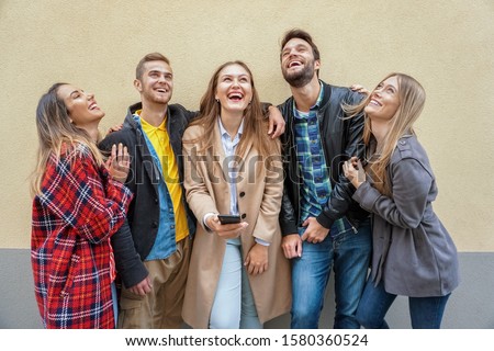Group of friends standing by the wall background and enjoying each other-  Teenagers using smartphone and smiling while trying to take a selfie- Technology concept