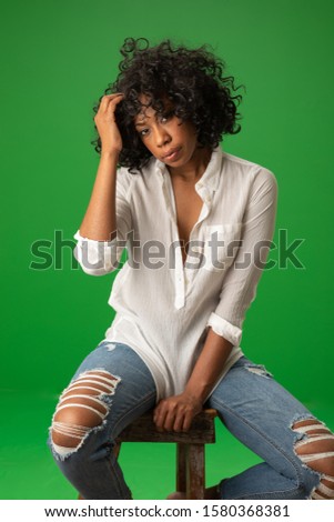 Portrait of pretty black female sitting on stool on greenscreen. Young woman wearing white blouse and looking at camera