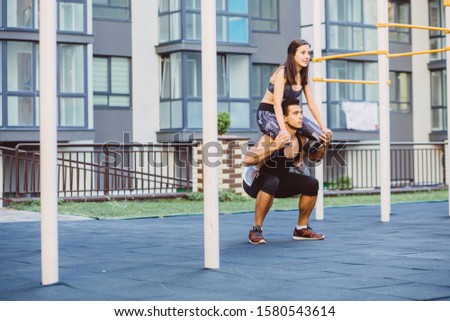 Mixed race couple is having fun on sportsground. Happy girl sitting on the shoulders of the guy squatting exercise, improvisation concept. Outdoor workout open air on urban background