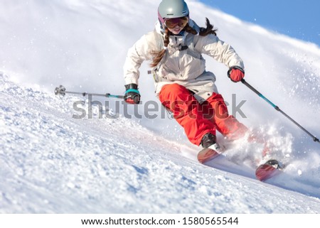 Girl On the Ski. a skier in a bright suit and outfit with long pigtails on her head rides on the track with swirls of fresh snow. Active winter holidays, skiing downhill in sunny day. Dynamic picture