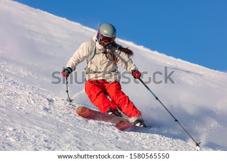 Girl On the Ski. a skier in a bright suit and outfit with long pigtails on her head rides on the track with swirls of fresh snow. Active winter holidays, skiing downhill in sunny day. Dynamic picture