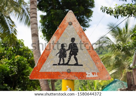 Old warning road sign depicting going children against the background of tropical trees