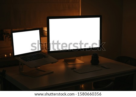 All-in-one desktop computer, laptop on the stand, two memory cards, wide-angle lens, keyboard, and mouse stands on the white table near the window with lamp and garland in the room where light off 