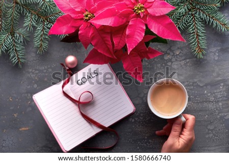 Christmas goals creative flat lay. Hand holding coffee, notebook with natural Xmas decorations, ribbon bookmark and pink trinket. Vibrant pink poinsettia plant and fir twigs on dark textured table.