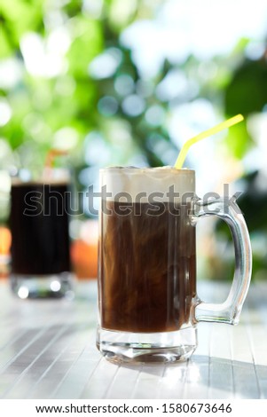 Subject shot of classic design beer mugs with dark refreshing drinks. The glass mugs with thick handles and coloured straws are situated on the white wood table in the summer outdoor interior.  
