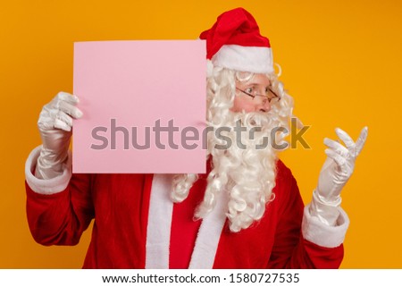 Santa Claus holds in his hands a pink sheet of paper for records and poses on a yellow background