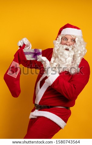 Santa Claus with a long beard and glasses holds a Christmas sock in his hands with gifts and boxes and poses on a yellow background