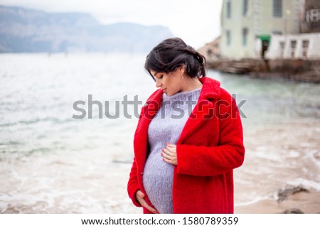 Pregnant woman walking at the beach at cold weather and dream about healthy maternity. female wearing red coat, warm sweater and jeans  at the background sea and mountain