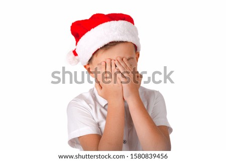 Close up Cute caucasian schoolboy with Christmas red hat and white t-shirt covers the face with hands. Picture white isolated.