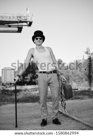 Young man, wearing grey pants, white top and black classic hat, holding old suitcase, leaning on cane. Black and white picture of creative man on abandoned construction site area. Art-house.