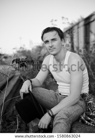 Young handsome man, wearing grey pants and white t-shirt, holding black classic hat, sitting on abandoned railway track by old train. Black and white picture of creative artist, posing for picture.