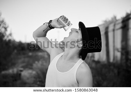 Close-up portrait of young man, wearing,white top, black classic hat, holding water glass, drinking last drops. Black and white picture of creative man on abandoned construction site.