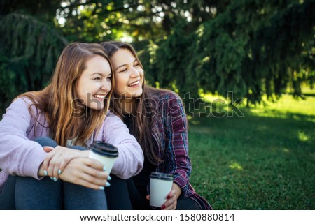 Attractive sisters drinking coffee on grass in park with copy space. Girls sit close to each other, look in one side and fun laughing. Homosexual relations, LGBT, family concept.