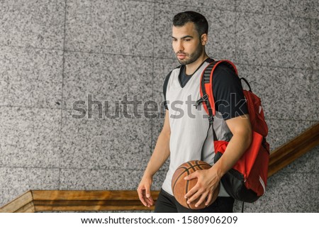 Attractive basketball player carries an orange ball and a red backpack, leaving the building where he lives