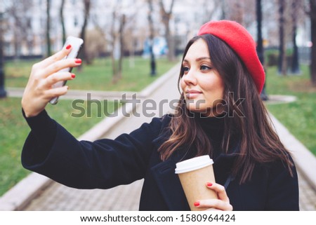 Beautiful young smiling brunette girl takes a selfie on the phone in autumn park in a black coat and red beret with a craft plastic cup with a hot drink in the afternoon.