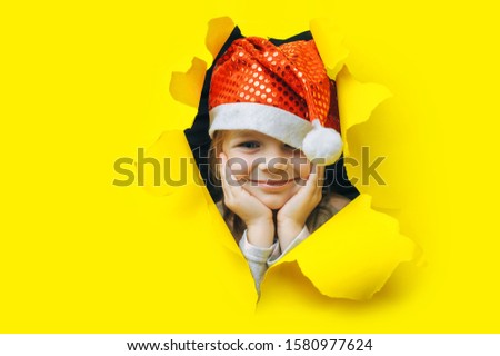 Waiting for a Christmas gift. A beautiful little girl in a red Santa hat put her head in her hands and looks out of a hole in yellow paper. Christmas and New Year concept. Copy space. Torn background.