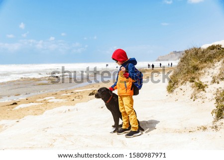 The boy stands with a dog at the seashore near the Scala dei Turchi in Sicilia. Boy kindly stroke his dog. The atmosphere is calm at the seashore. White sand makes the picture light