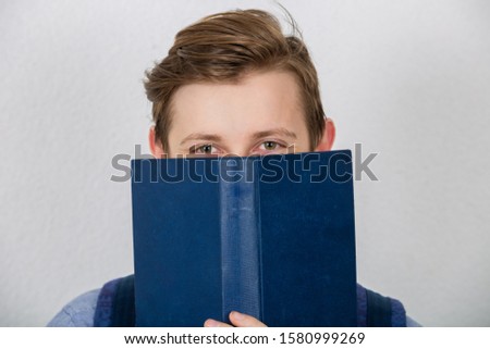  Happy teenager boy smiling covering half face with a opened blue book. Book lover. Ready to study hard.