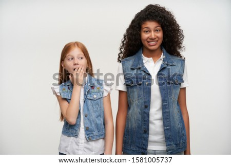 Bewildered young curly brunette woman with dark skin looking confusedly to camera and raising eyebrows while posing over white background with amazed red haired little girl