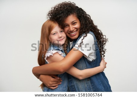 Portrait of attractive happy young dark skinned woman with long curly hair hugging pretty red haired little girl and smiling widely while posing over white background
