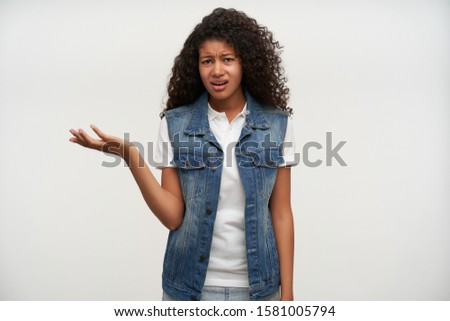 Displeased young dark haired curly woman with dark skin pouting and frowning eyebrows with raised hand while standing over white background, dressed in casual clothes