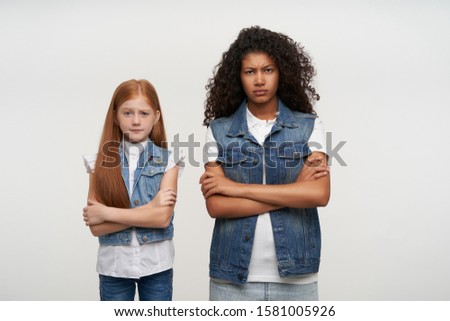 Severe young ladies with long hair standing over white background with folded hands, looking severely at camera and frowning eyebrows, dressed in casual family look