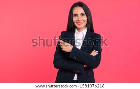 Look what I did! Close-up photo of a stunning young woman in a smart outfit, who is posing with folded arms, smiling at the camera and pointing to the left with her right index finger.