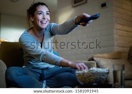 Young happy woman changing channels with remote control while watching TV and eating popcorn in the evening at home.  Royalty-Free Stock Photo #1581091945
