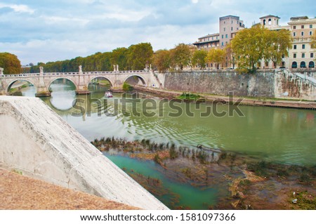 Image of Rione Ponte district. View of white Ponte Sant'Angelo bridge and its reflection, orange trees, green waters of Tiber river under dramatic cloudy autumn sky