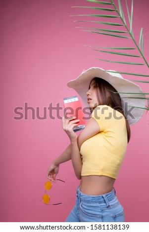 Beautiful girl in a huge white hat with a passport in her hands in glasses is going on a trip and rejoices in a yellow T-shirt and pink background
