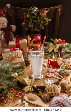 Christmas and New Year eggnog drink in glass cup on a wooden stand on the table with gifts, Christmas cookies, candles, sweets and Christmas tree branches. for holiday drink recipes.