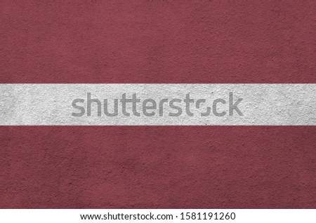 Latvia flag depicted in bright paint colors on old relief plastering wall. Textured banner on rough background