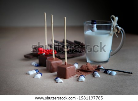 Small snowman near Chocolate cubes on a stick and cacao spoon with milk cup and aromatic cocoa and chocolate on brown background, close up. Hot cocoa with milk in transparent glass cup, broken chocola
