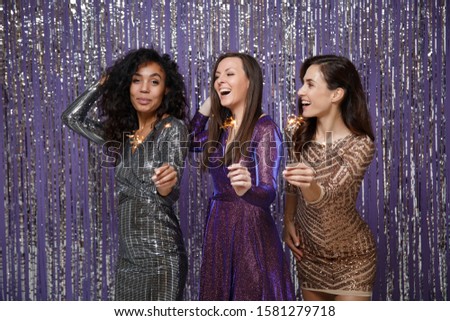 Women in fancy sparkling dresses posing isolated over vibrant purple violet silver background. Positive people spending time on happy birthday new year holiday party discotheque hold burning sparkler