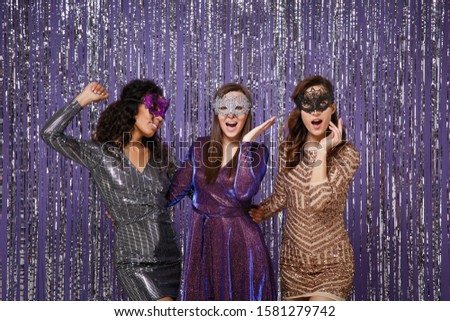 Beautiful mysterious women in fancy sparkling dresses carnival masks posing isolated over vibrant purple violet silver background. People spending time on happy birthday new year holiday party disco