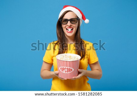 Cheerful woman Santa girl in Christmas hat 3d imax glasses posing isolated on blue background. New Year 2020 celebration holiday concept. Mock up copy space. Watch movie film hold bucket of popcorn