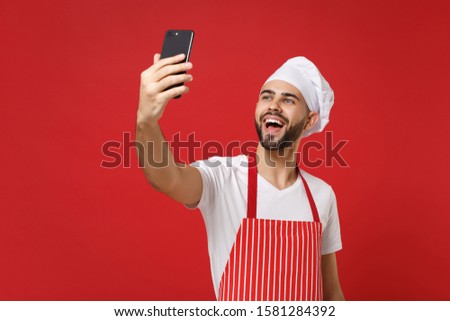 Cheerful young bearded male chef cook or baker man in striped apron toque chefs hat isolated on bright red wall background. Cooking food concept. Mock up copy space. Doing selfie shot on mobile phone